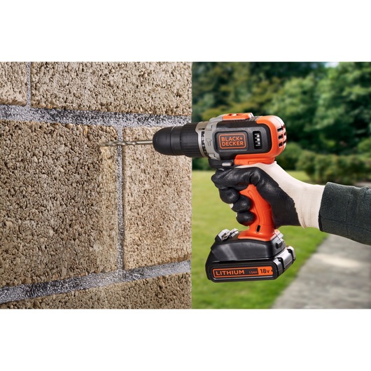 A person wearing gloves uses a BLACK+DECKER cordless drill to drill into a concrete block wall outdoors. The drill is orange and black and labeled with 'BLACK+DECKER' and 'LITHIUM 18V.'