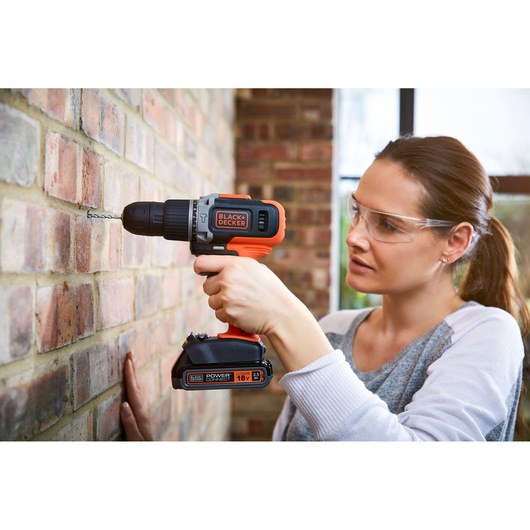 A person using a BLACK+DECKER cordless drill to drill into a brick wall. The individual’s face is blurred for privacy. The drill is orange and black, and the scene is set indoors.