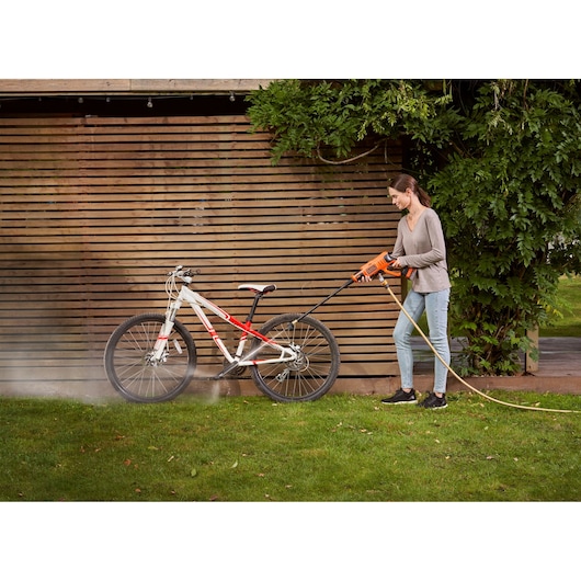 A person is using a BLACK+DECKER pressure washer to clean a bicycle outdoors in a garden area, with a wooden fence and greenery in the background.