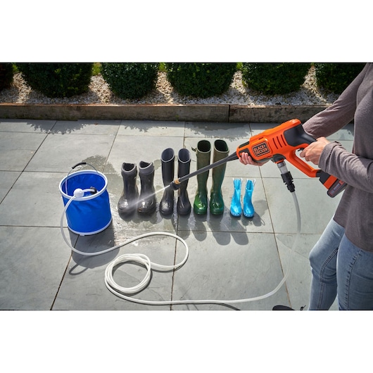 A person uses a BLACK+DECKER portable pressure washer to clean boots arranged in a row outside on patio tiles. A blue bucket with a hose is used to supply water.