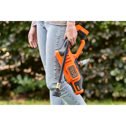 A person holding a BLACK+DECKER portable pressure washer outdoors. The orange and black device is being carried by the handle, with a hose attached, against a natural green background.