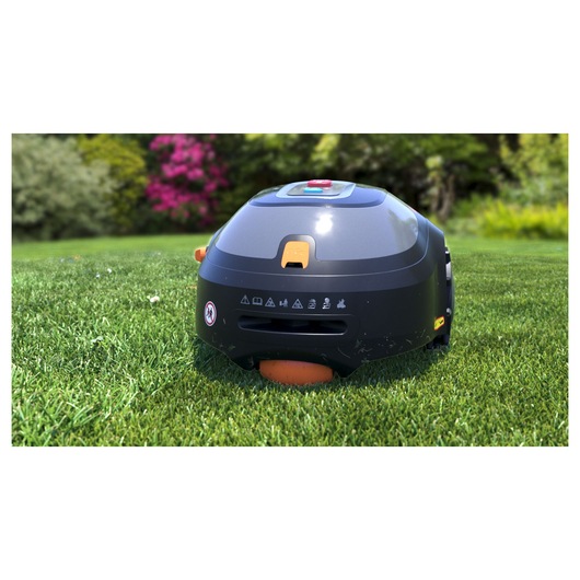 A BLACK+DECKER robotic lawn mower (model BCRMW121) is shown on a green lawn outdoors, with trees and flowers in the background. The device is black with orange accents and has warning icons on its front.