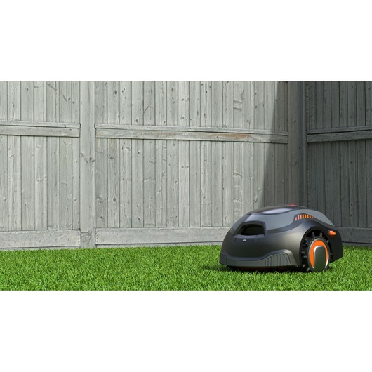 A BLACK+DECKER robotic lawn mower is positioned on grass near a wooden fence in an outdoor yard.