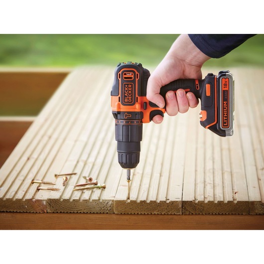 A hand holding a BLACK+DECKER cordless lithium 18V drill, screwing into a wooden surface with several screws lying nearby.