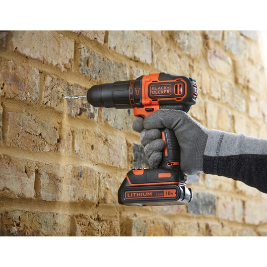 A person wearing gray gloves is using a BLACK+DECKER cordless drill with an orange and black design to drill into a brick wall.