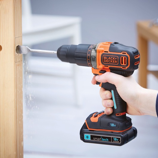 A hand holding a BLACK+DECKER cordless drill with an 18V lithium battery, drilling a hole into a wooden surface.