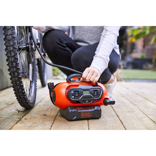 A person is using a BLACK+DECKER portable air compressor to inflate a bicycle tire on a wooden deck.