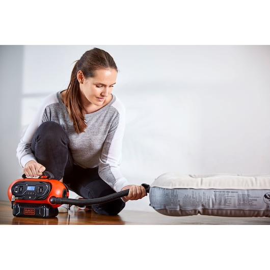 A person is kneeling on the floor and using a BLACK+DECKER BDCINF18N inflator to inflate an air mattress. The inflator is orange and black, with a digital display and attached hose. The person's face is blurred for privacy.