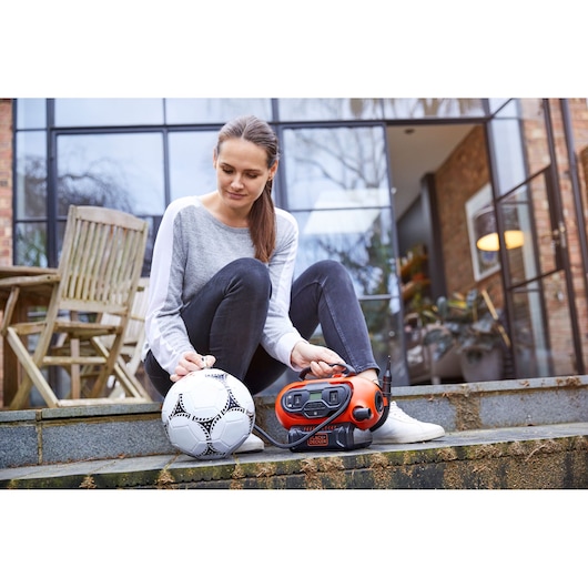 A person sitting on outdoor steps next to a BLACK+DECKER BDCINF18N inflator and a soccer ball, preparing to inflate the ball. The product is orange and black, positioned near the person's foot. The background shows a wooden chair and a modern glass door.