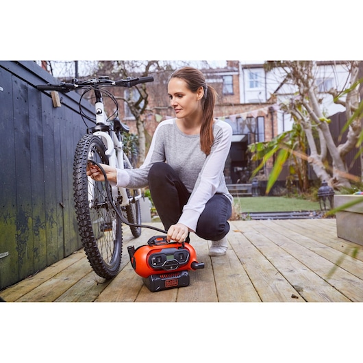 A person using a BLACK+DECKER portable air compressor (BDCINF18N) to inflate the tire of a bicycle outdoors on a wooden deck. The scene shows a modern garden setting.
