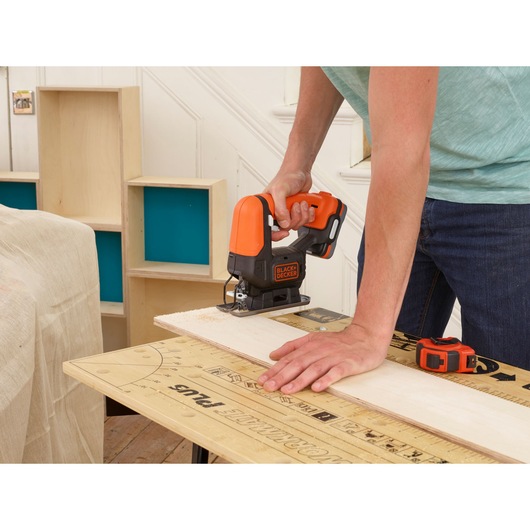BLACKDECKER BDCK123S2S cordless jigsaw cutting wood on workbench, orange and black tool in use.