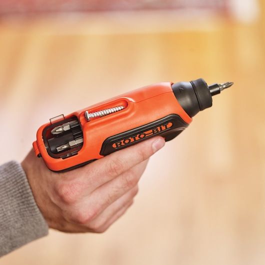 A close-up image of a hand holding an orange and black BLACK+DECKER electric screwdriver with visible screwdriver bits and a screw stored in the built-in compartment.