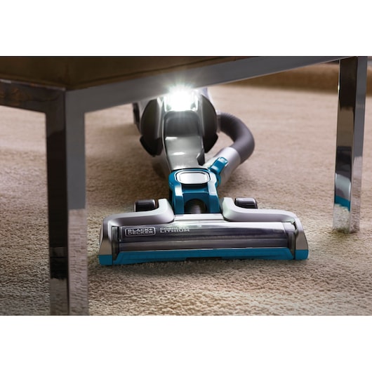 A BLACK+DECKER cordless vacuum cleaner with a lithium battery cleaning carpet under a piece of furniture, with an LED light illuminating the area under the furniture.