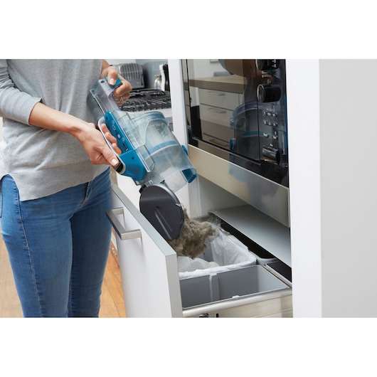 A person is emptying the dust container of a cordless vacuum cleaner into a kitchen trash bin.