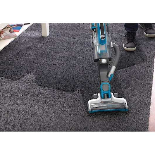 A BLACK+DECKER cordless vacuum cleaner being used to clean a dark carpet, with part of a person's legs visible and a white table with magazines in the background.