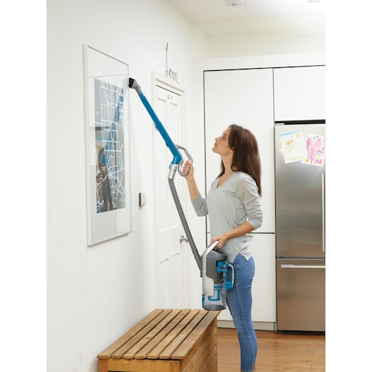 A person is using a BLACK+DECKER CUA525BH cordless vacuum cleaner with a long attachment to clean the top of a framed picture on a white wall in a modern kitchen.