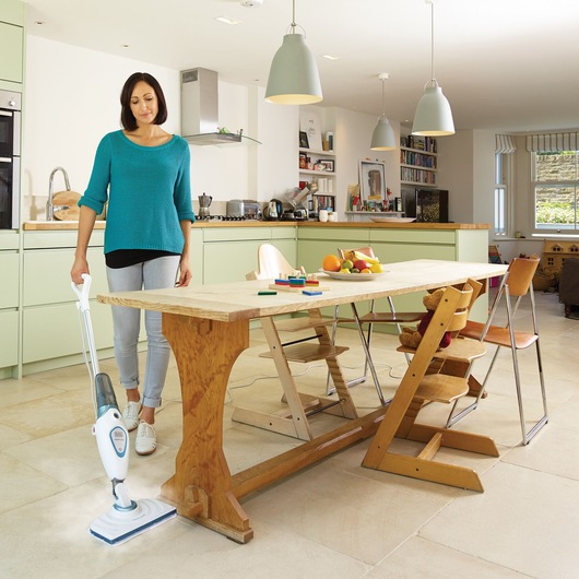 A person using a BLACK+DECKER FSM1605 steam mop to clean the floor in a modern kitchen and dining area. There is a wooden dining table with fruit and toys on top, surrounded by various chairs. The kitchen is equipped with light green cabinets, modern appliances, and pendant lights.