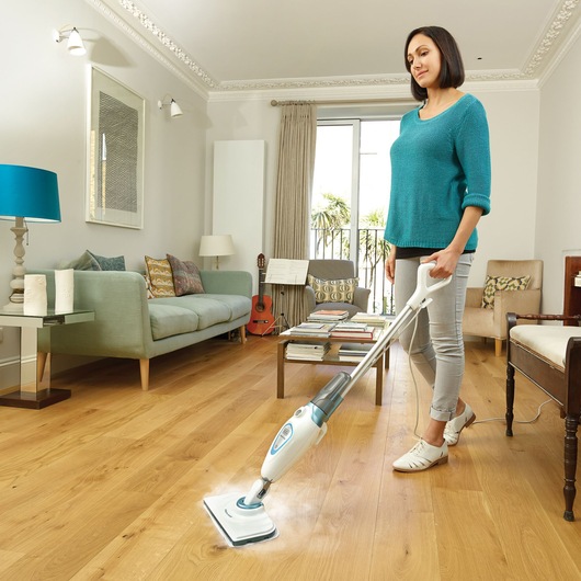 A person is using a BLACK+DECKER FSM1605 steam mop to clean a wooden floor in a modern living room. The room is bright and includes a sofa, guitar, table with books, and large windows with curtains.