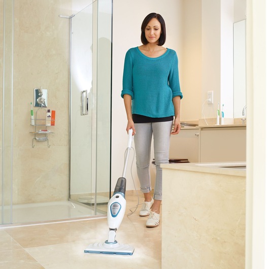 A person cleaning the bathroom floor with a BLACK+DECKER steam mop. The room features a shower, sink, and beige tiled surfaces.