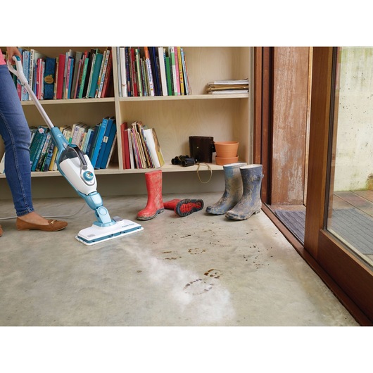 A person is cleaning a dirty concrete floor with a BLACK+DECKER steam mop, leaving clean streaks where muddy footprints are. In the background, there is a bookshelf filled with books and some muddy boots placed on the floor near the door.