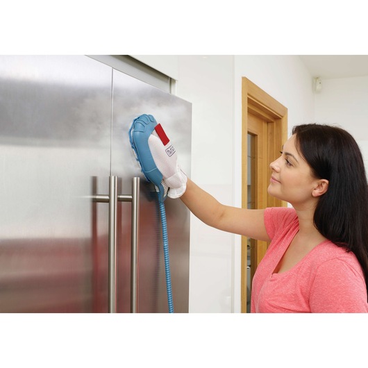 A person uses a BLACK+DECKER steam cleaning glove to clean a stainless steel refrigerator door in a modern kitchen. The person's face is blurred for privacy.