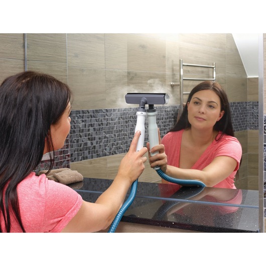 A person using a BLACK+DECKER steam handheld cleaner to clean a bathroom mirror. The device emits steam, and the person is holding the cleaner with both hands while wearing a pink shirt. The bathroom has tiled walls and a towel on the counter.