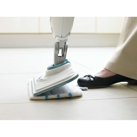 A close-up of a person using a BLACK+DECKER steam mop on a tiled floor. The person's foot is pressing a button on the mop, and the mop head is equipped with a cleaning pad.