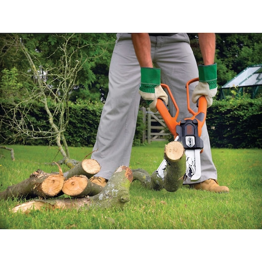 A person wearing gloves and protective clothing uses an orange and black BLACK+DECKER chainsaw to cut tree branches into smaller pieces on a grassy lawn.