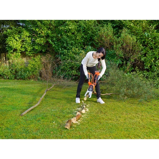 A person using a BLACK+DECKER GKC1000L cordless chainsaw to cut branches on a lawn, surrounded by green bushes and trees in a garden.
