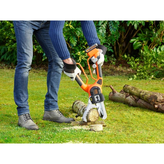 A person using a BLACK+DECKER cordless chainsaw (model GKC1000L) to cut a small log outdoors on grass, wearing gloves and casual clothing. The log is placed on the ground with other logs in the background.