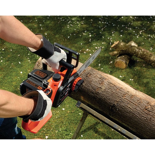 A person wearing gloves is using a BLACK+DECKER cordless chainsaw to cut a log placed on a metal support in a grassy outdoor setting.