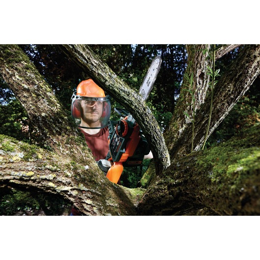 Person wearing protective gear using a BLACK+DECKER chainsaw to cut tree branches outdoors. The chainsaw model is GKC3630L20.