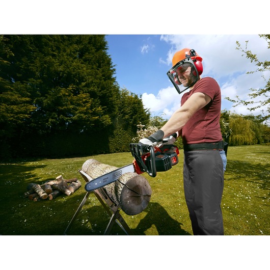 A person wearing protective gear is using a BLACK+DECKER cordless chainsaw outdoors to cut a large log placed on a sawhorse. Other logs are visible in the background, and the scene is set in a garden with green grass and trees.