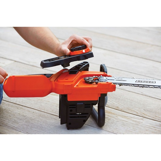 A person is performing maintenance on an orange and black cordless chainsaw, with the cover of the chain mechanism being lifted to show the internal parts. The chainsaw is placed on a wooden surface.