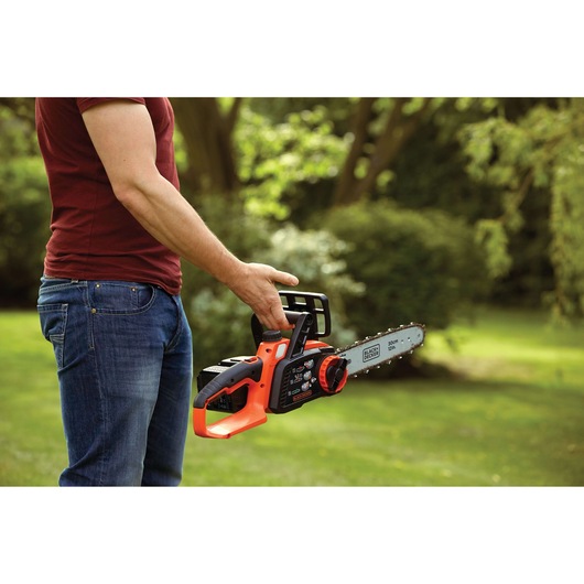 A person holding a BLACK+DECKER cordless chainsaw outdoors on a grassy area, with trees and shrubs in the background.