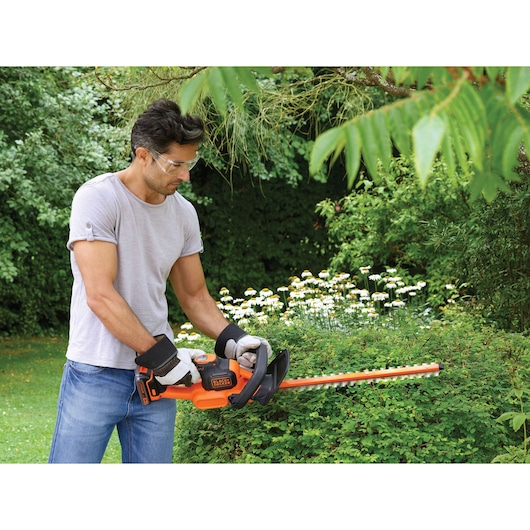 A person with a blurred face is using a BLACK+DECKER cordless hedge trimmer to trim bushes in a garden. The trimmer is orange and black, and the person is wearing gloves and a grey T-shirt.