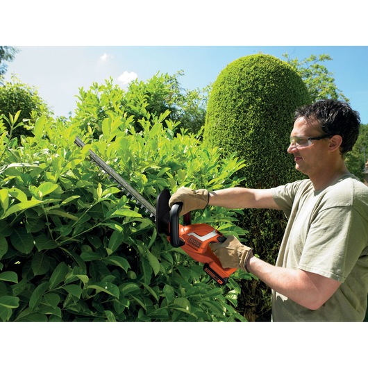 A person with a blurred face is using a BLACK+DECKER orange cordless hedge trimmer to trim green bushes in a garden, wearing gloves and a t-shirt.