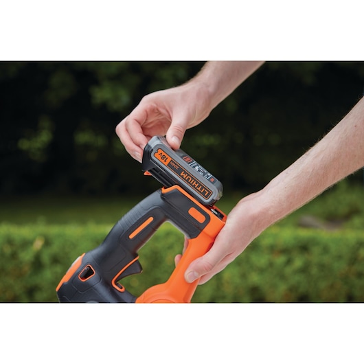 A close-up of a person inserting an 18V lithium battery into a BLACK+DECKER cordless power tool outdoors, with green foliage in the background.