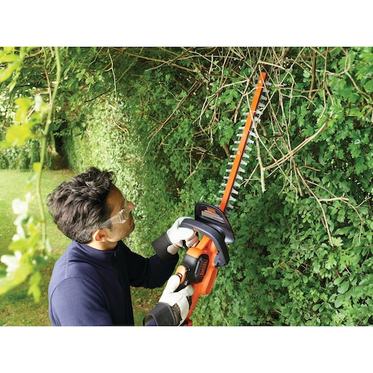 A person wearing gloves uses a BLACK+DECKER GTC18502PC hedge trimmer to trim branches and foliage outdoors. The person's face is blurred for privacy.