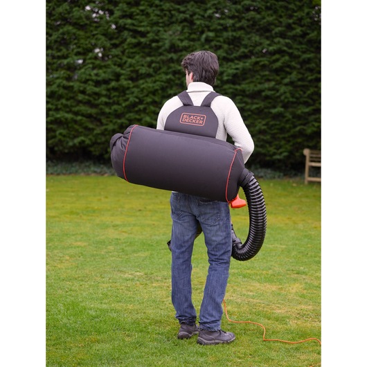 A person outdoors wearing a BLACK+DECKER branded photography equipment backpack with a large cylindrical shape and black hose attachment, standing on grass near a hedge.