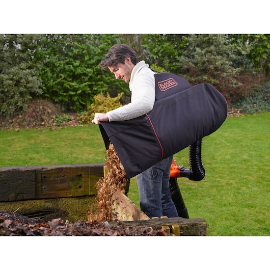 A person using a BLACK+DECKER garden vacuum with a large collection bag, emptying leaves into a wooden compost area in a garden. The BLACK+DECKER logo is visible on the bag.