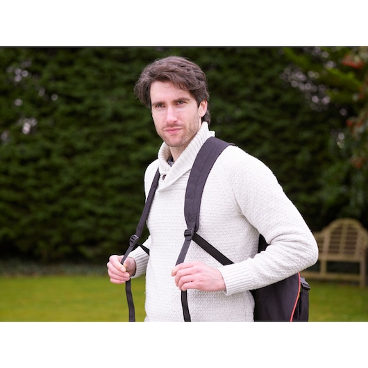 A person wearing a light-colored sweater and carrying a black backpack is standing outdoors, with greenery and a bench in the background. The face is blurred for privacy.
