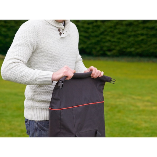 A person wearing a light-colored sweater is holding a BLACK+DECKER black bag with red trim, standing outdoors on a grassy area.