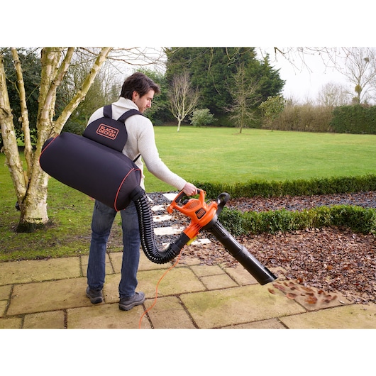 A person using a BLACK+DECKER backpack leaf blower on a patio to clear fallen leaves. The person is wearing the black and orange branded backpack, connected to the orange blower, in a garden with grass and trees.