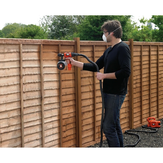A person using a BLACK+DECKER HVLP400 paint sprayer to paint a wooden fence outdoors. The sprayer is connected to a hose and a base unit placed on the ground.