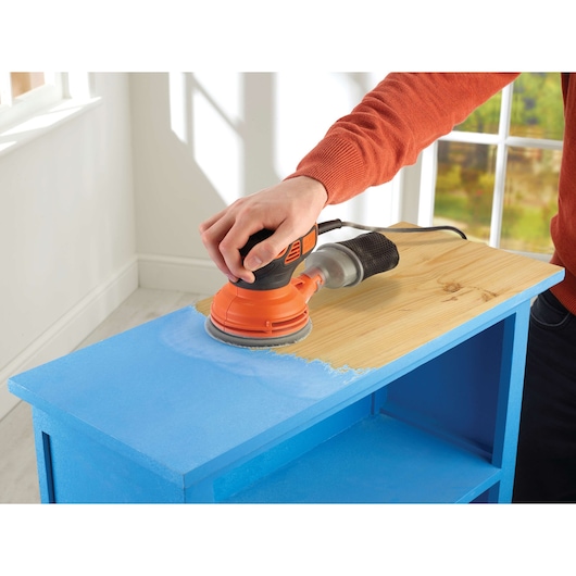 A person is using a BLACK+DECKER KA199 orange electric sander to remove blue paint from the top surface of a wooden cabinet near a window.
