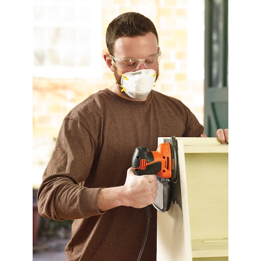 A person using a BLACK+DECKER KA2500K electric sander on a wooden piece of furniture. The person is wearing a brown long-sleeve shirt and safety goggles. Their face is not visible.