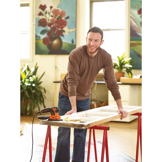 A person with a blurred face is working indoors on a wooden surface, using a BLACK+DECKER KA2500K power tool placed on a table supported by red sawhorses. Art and plants decorate the background.