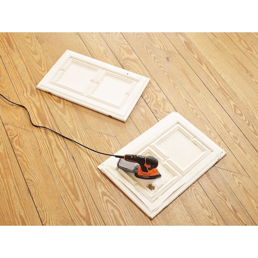 A BLACK+DECKER KA2500K electric sander placed on a white wooden cabinet door, next to another similar door, both lying on a light wood floor. The sander is plugged in, ready for use.