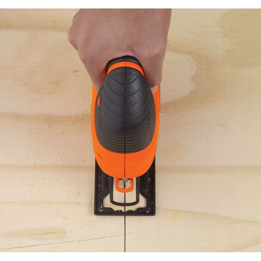 A hand holding an orange and black BLACK+DECKER jigsaw, cutting along a straight line on a wooden board.